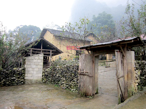 Không gian ngôi nhà nhìn từ ngoài cổng với tường trình, cửa gỗ, hiên đá, mái lợp ngói máng